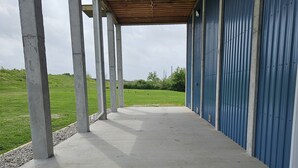 Een overdekt buitengebied met betonvloer en metalen balken, dat leidt naar een blauw gebouw met een golfplaten gevel.
