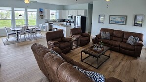 Een ruime woonkamer met leren banken, een salontafel en een eethoek met een tafel en stoelen. Er zijn plafondventilatoren en een keuken zichtbaar op de achtergrond.