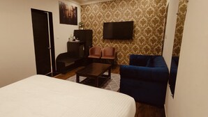 Quarto de hotel com cama, escrivaninha, duas cadeiras, televisão e parede estampada.