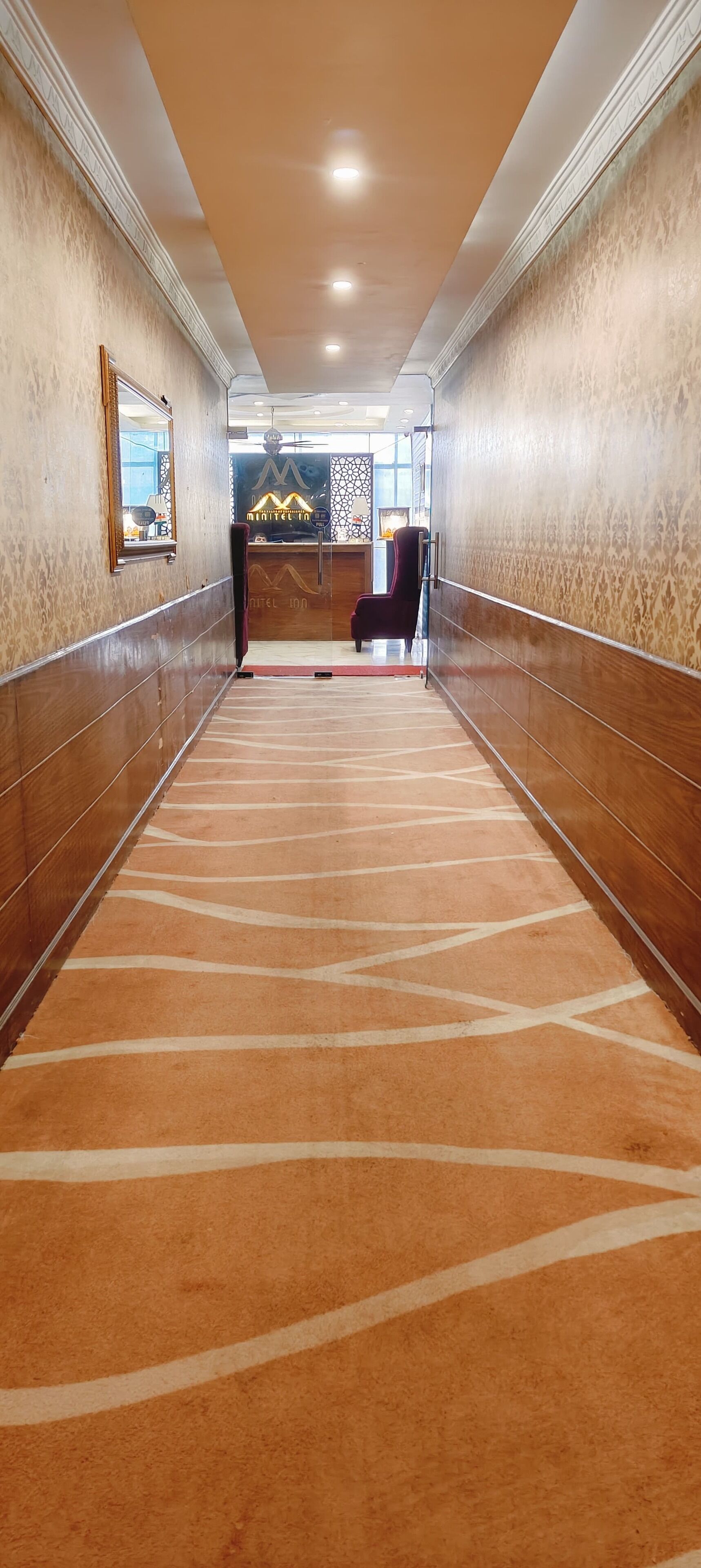 A hotel corridor with patterned carpet, wooden paneling, and a reception desk in the distance.