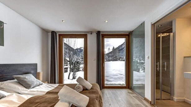 A modern bedroom with a large bed, two windows with a snowy mountain view, and a sliding glass door leading to a bathroom. - Chalet 200m2 | Fireplace | Jacuzzi | Sauna | Views (Chamonix-Mont-Blanc)