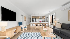 Ein modernes Wohnzimmer mit einem Flachbildfernseher, einer Couch, einem Essbereich mit einem Holztisch und einer Küche im Hintergrund.