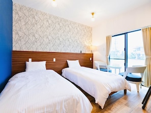 A hotel room with two beds, a patterned wall, a window with curtains, a small table, and a chair.