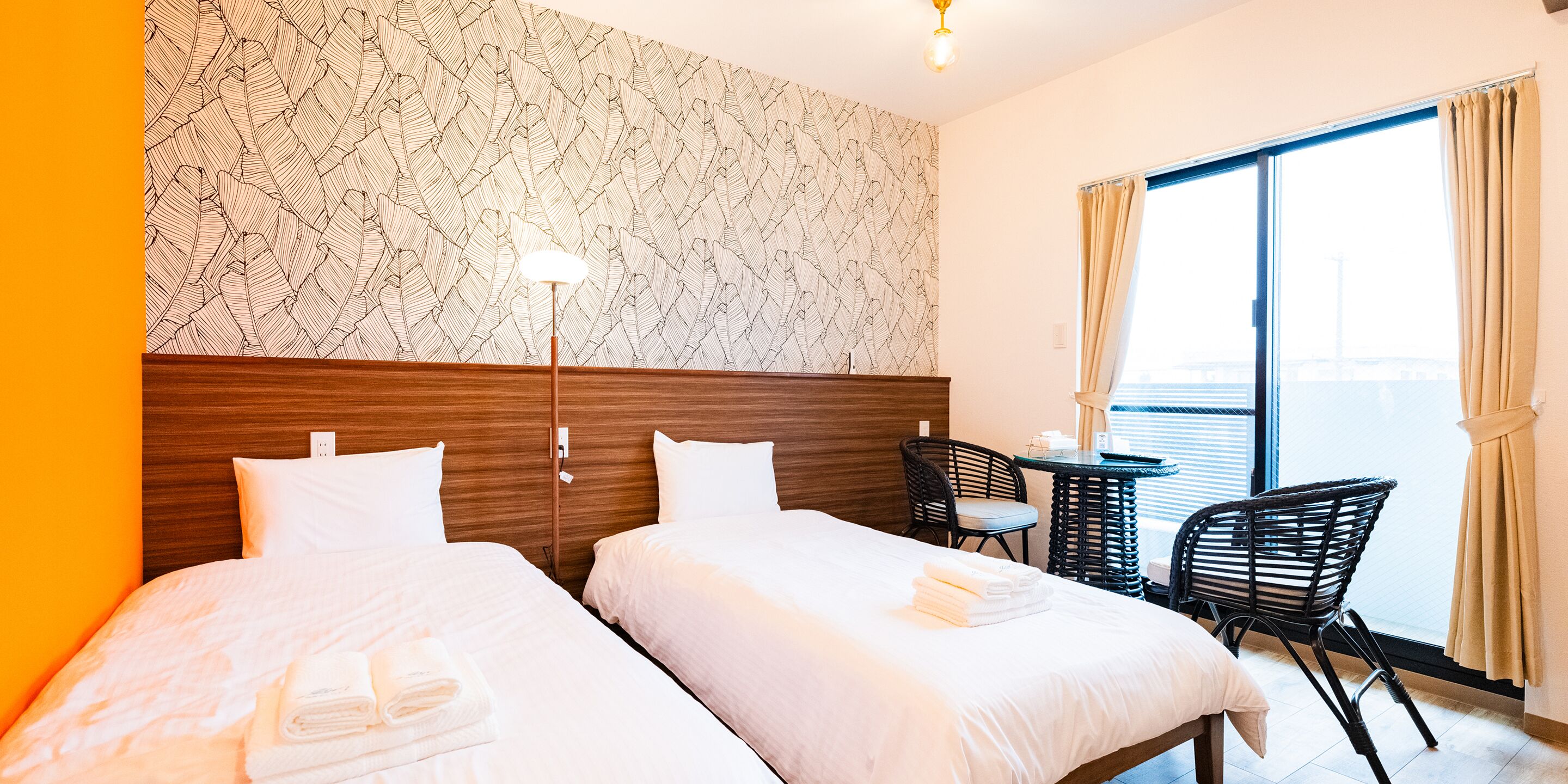 Quarto de hotel com duas camas, cabeceira de madeira, uma pequena mesa com uma cadeira e vista da cidade através de grandes janelas.