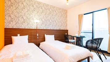 Quarto de hotel com duas camas, cabeceira de madeira, uma pequena mesa com uma cadeira e vista da cidade através de grandes janelas.