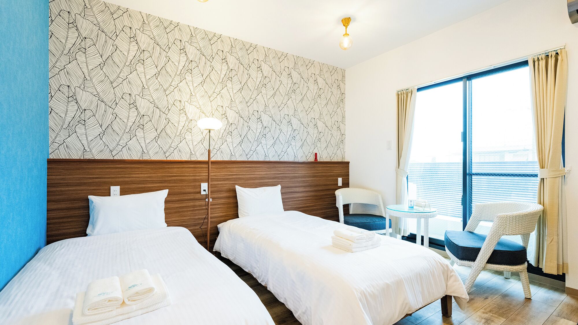 A hotel room with two beds, a small table, a chair, a floor lamp, and a large window with curtains.