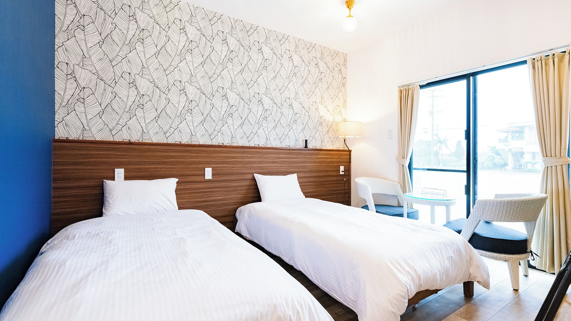A hotel room with two beds, a large window with curtains, a small table, and a chair.