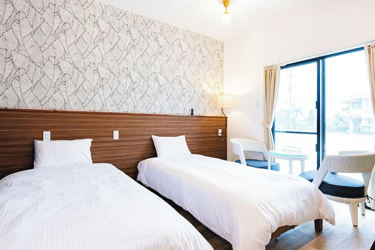 Una habitación de hotel con dos camas, un ventanal con cortinas, una mesita y una silla.