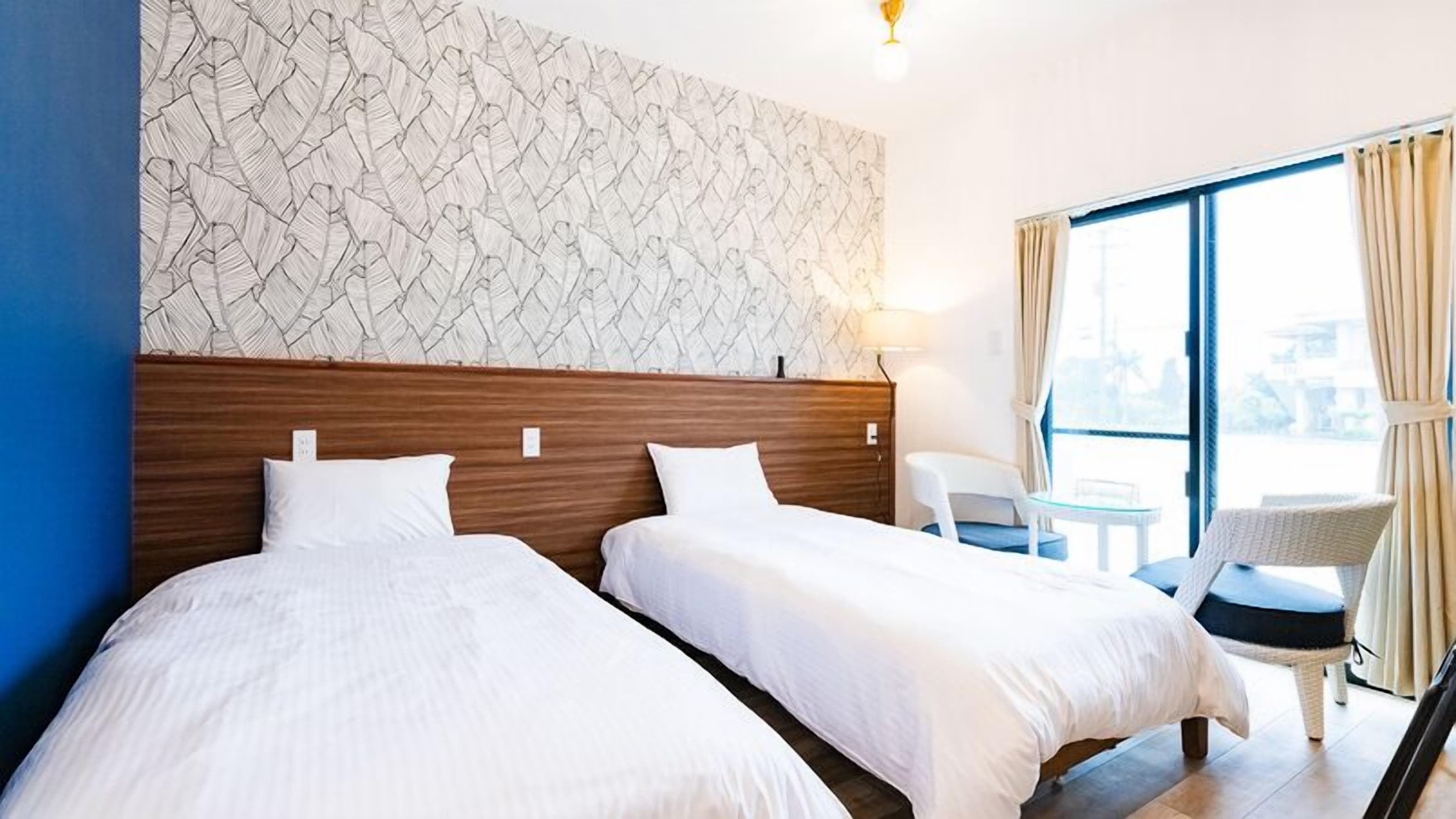 Habitación de hotel con dos camas, cabecero de madera, pared con diseño, ventana con cortinas y una pequeña mesa con una silla.