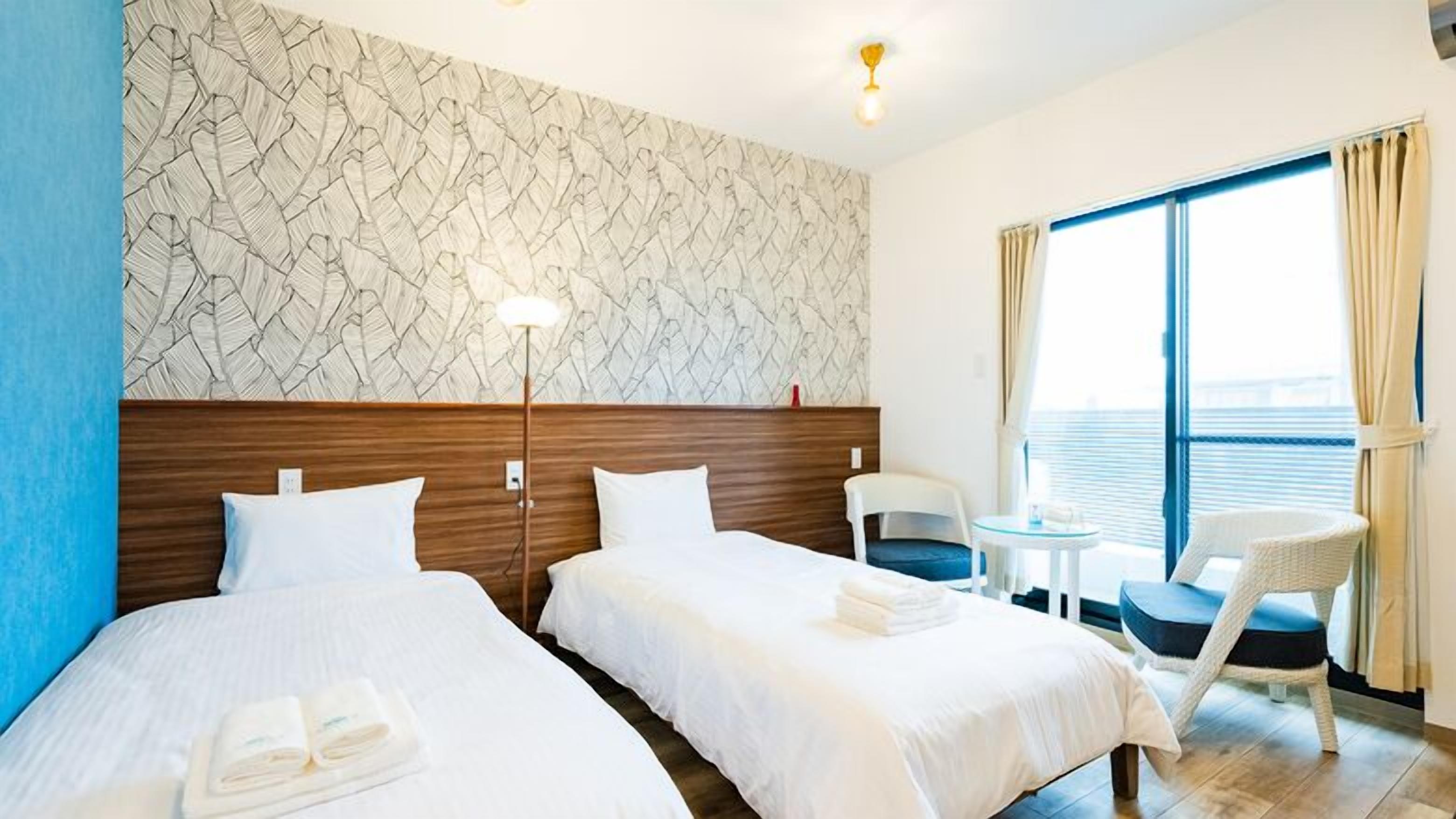 Una habitación de hotel con dos camas, una mesita, una silla, un ventanal grande con cortinas y una pared con estampado.