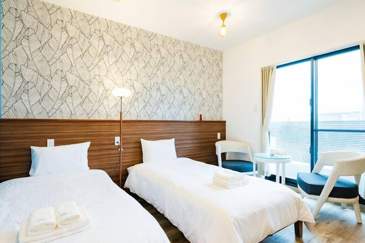 Une chambre d’hôtel avec deux lits, une petite table, une chaise, une grande fenêtre avec des rideaux et un mur à motifs.