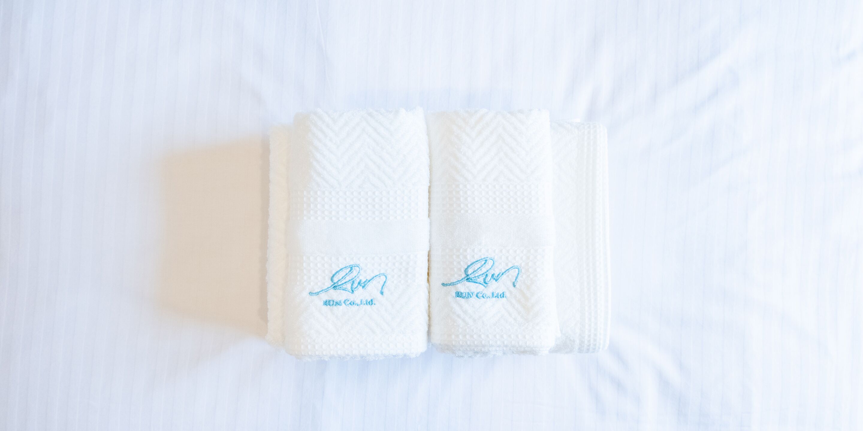 Deux serviettes blanches, soigneusement pliées, ornées d’un motif brodé, reposent sur un couvre-lit blanc.