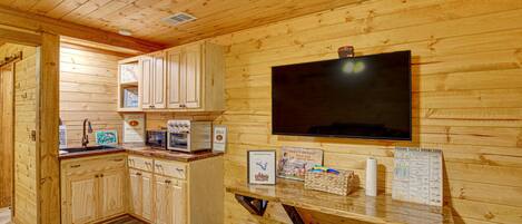 A wooden cabin interior with a kitchen area, wooden cabinets, a microwave, a sink, a television mounted on the wall, and a wooden bench.