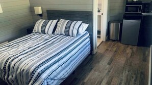 Un dormitorio con una cama, lámparas de noche, un cabecero de madera, una puerta que da a una cocina y un microondas.