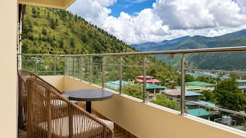 A balcony with a wicker chair and a small table, overlooking a mountainous landscape with houses.