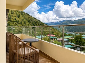 A balcony with a wicker chair and a small table, overlooking a mountainous landscape with houses. - Thimphu Deluxe (Thimphu)
