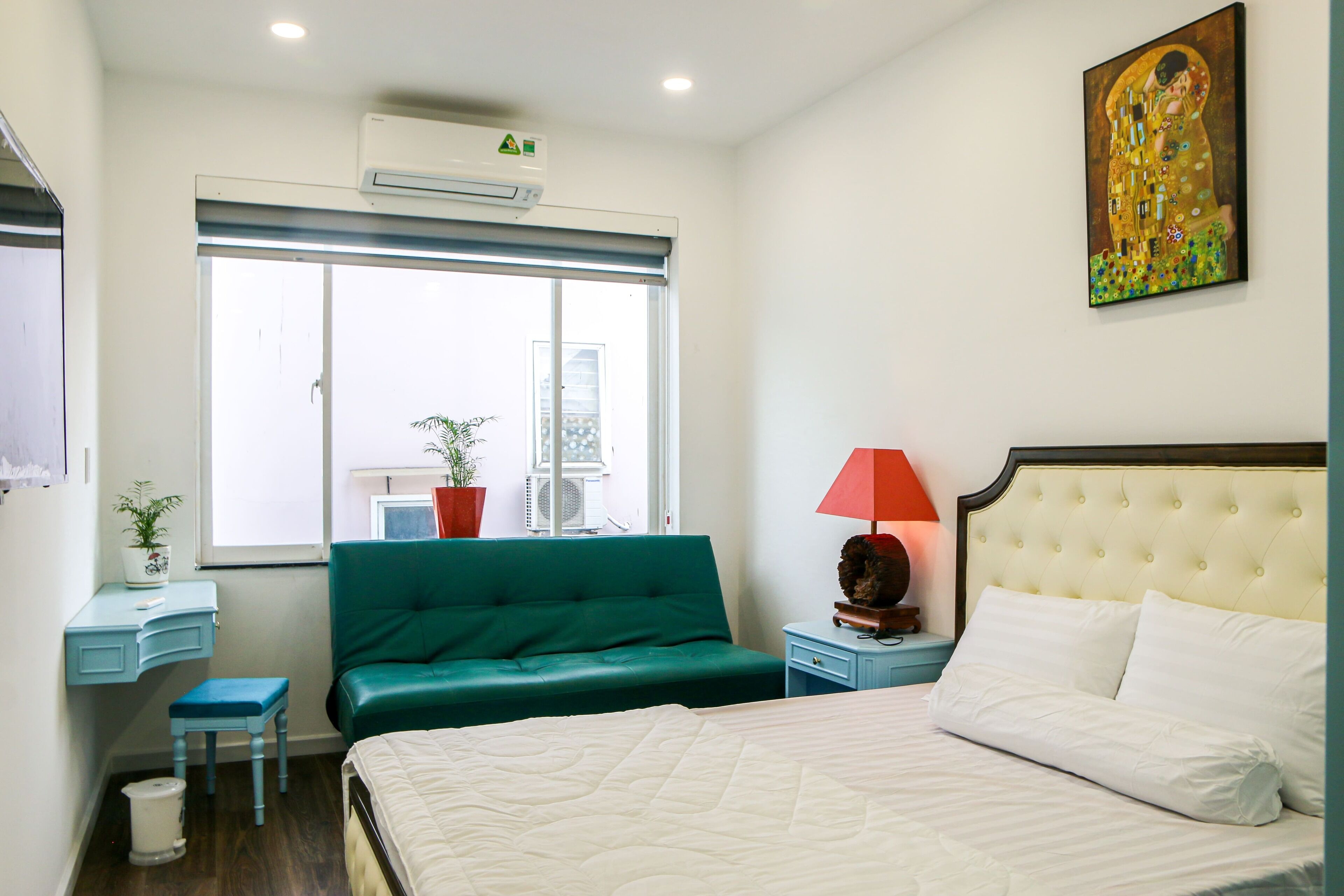 A bedroom with a bed, a green sofa, a desk with a chair, a window, and a painting on the wall.