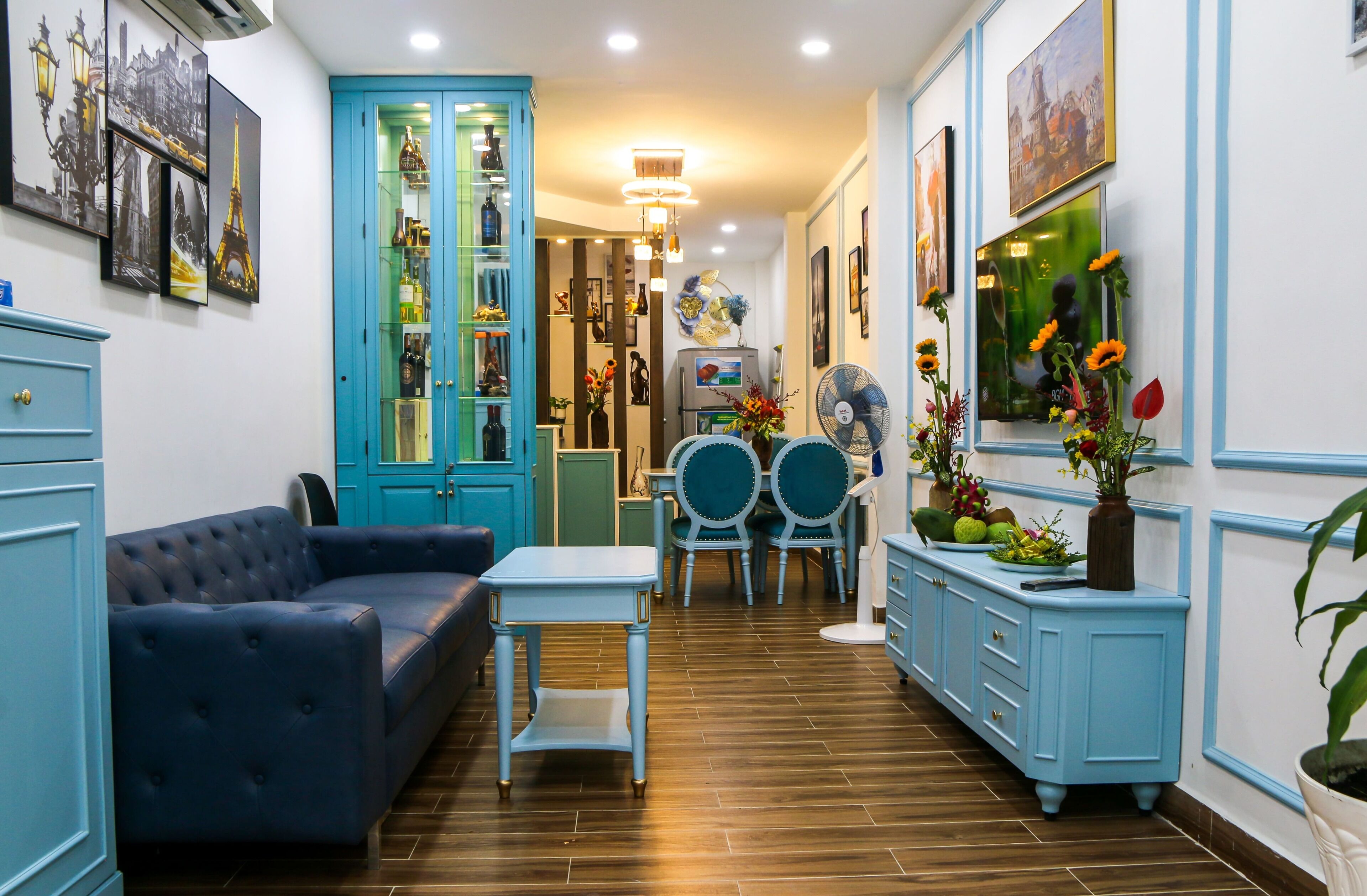 Una habitación con un sofá azul, una mesita pequeña, un armario azul y una zona de comedor con dos sillas y un ventilador.