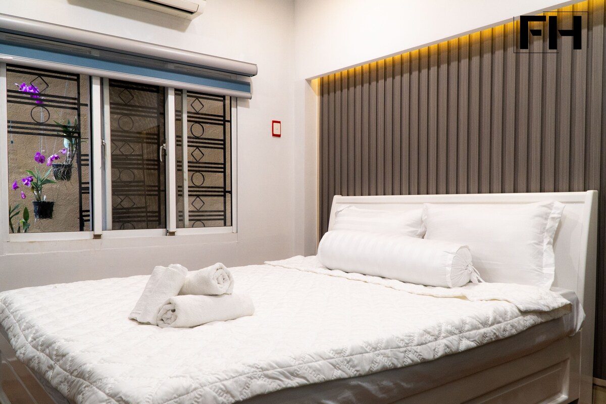 A neatly made bed with white linens and pillows, a window with a view of a building, and a wall with vertical lines and a light fixture.