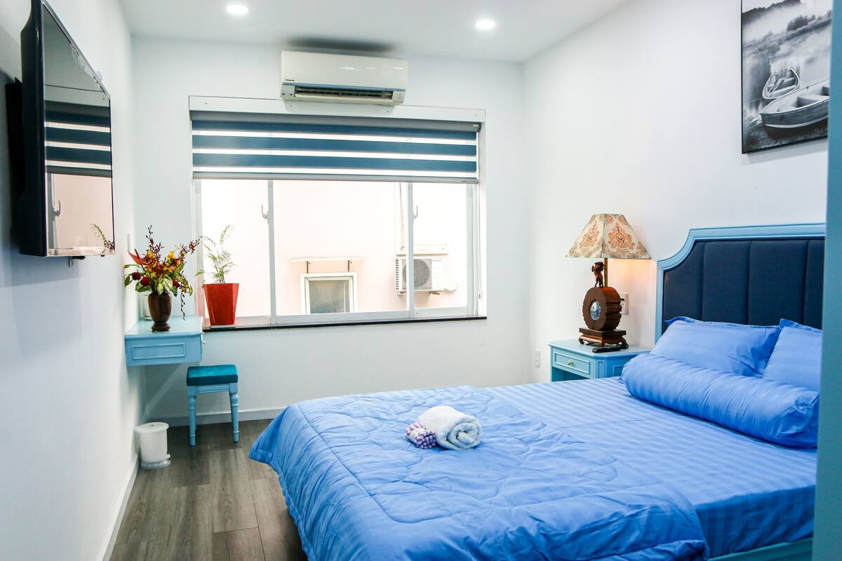 Une chambre à coucher comprenant un lit, un bureau, une chaise, une fenêtre avec des stores, un climatiseur fixé au mur et une lampe.
