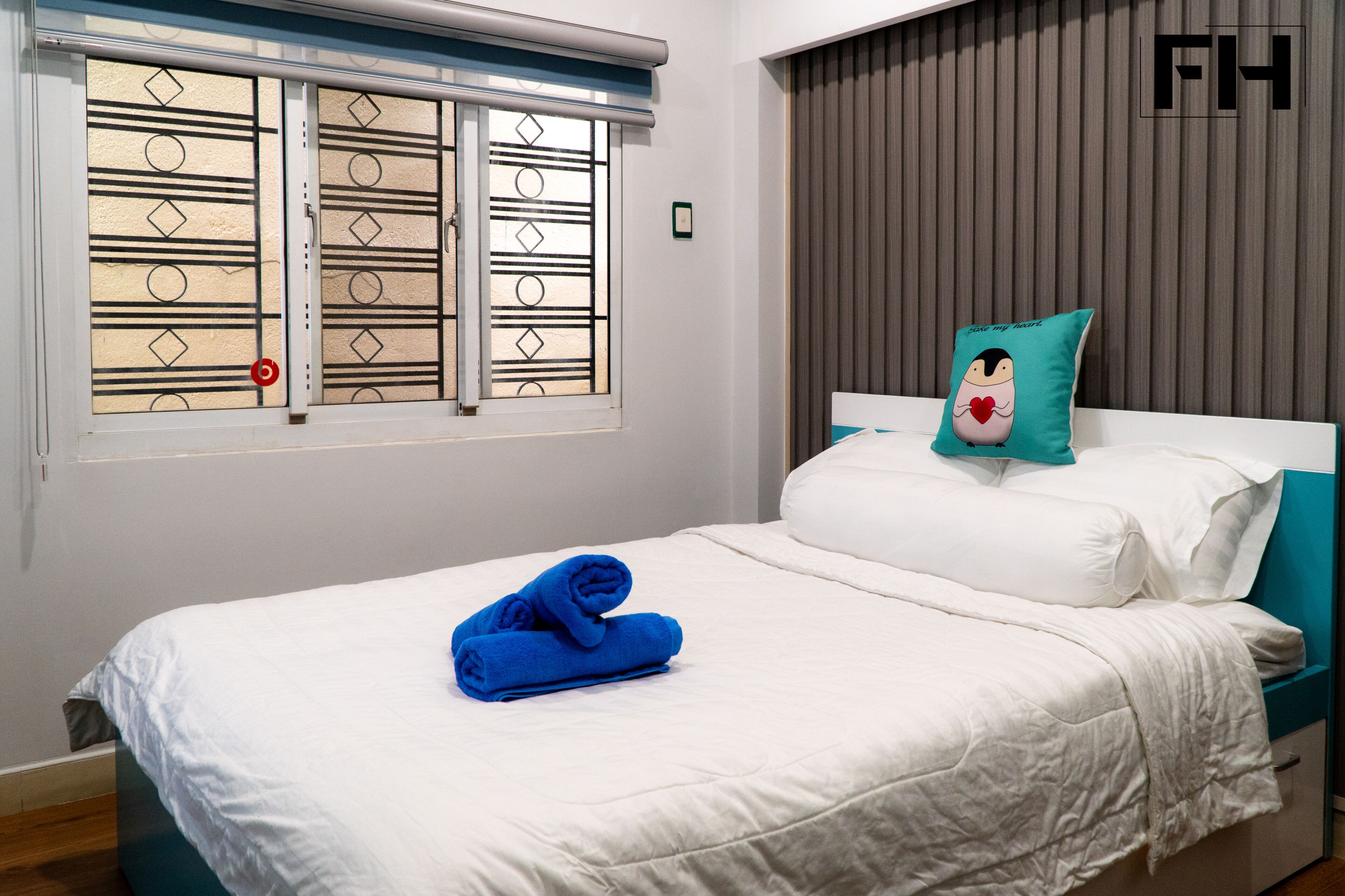 A neatly made bed with white and teal bedding, a teal pillow with a cartoon character, and a window with decorative blinds.