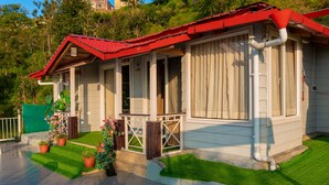 Ein eingeschossiges Haus mit rotem Dach, einer Veranda mit Geländer und einem kleinen Garten mit Blumentöpfen und Blumen.