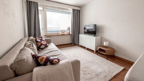 Una sala de estar moderna con un sofá, un televisor, una mesita auxiliar y un ventanal con cortinas.