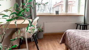 Una habitación con escritorio de madera, cama con cubrecama acolchado, planta en maceta y vista a edificios a través de una ventana.