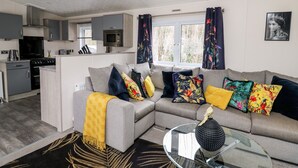 A modern living room with a grey sofa, colorful cushions, a glass coffee table, and a kitchen area with grey cabinets and a microwave. - The Hideout (New Quay)