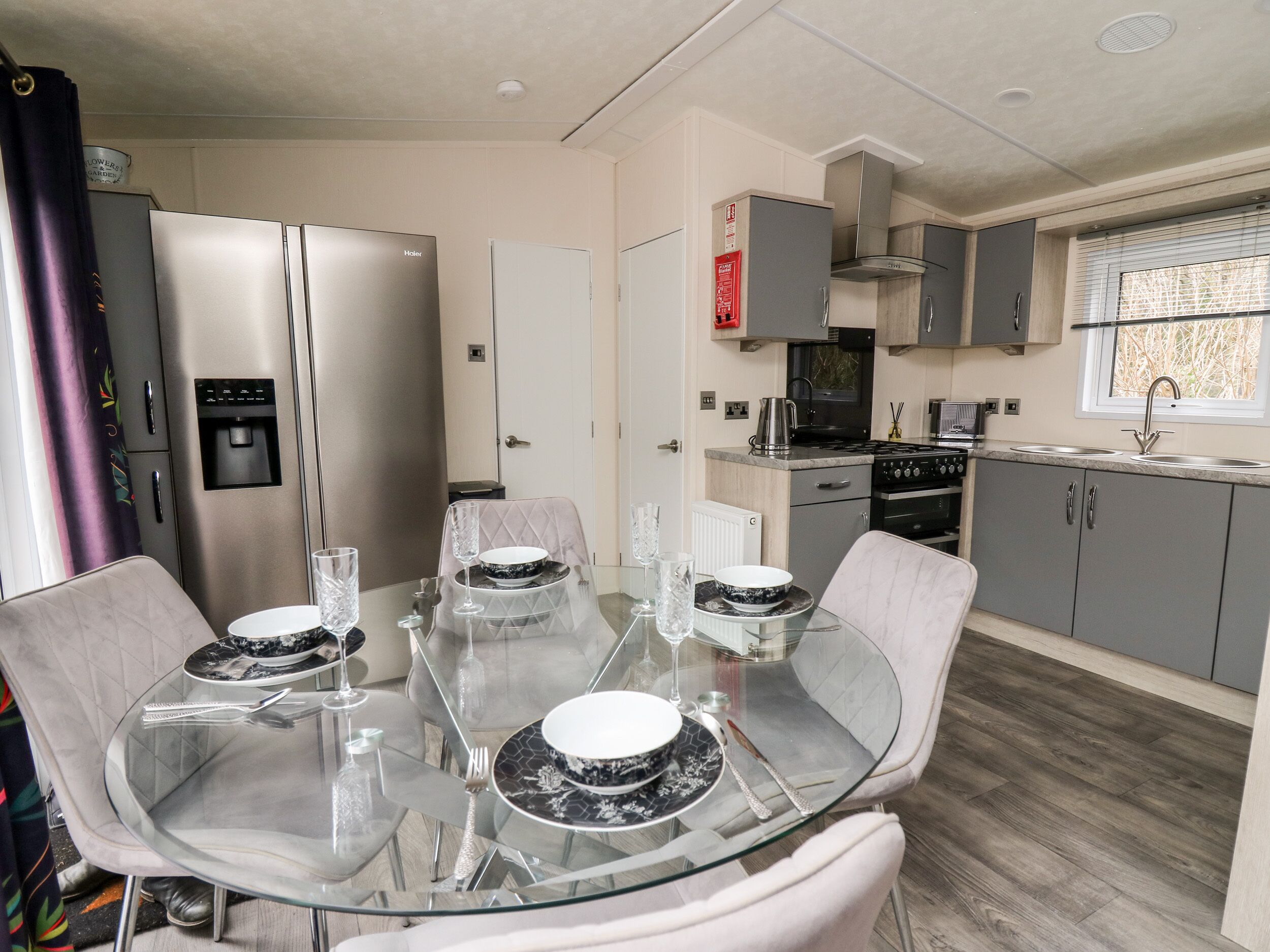 A modern kitchen with grey cabinets, a stainless steel refrigerator, and a dining area with a glass table and chairs.