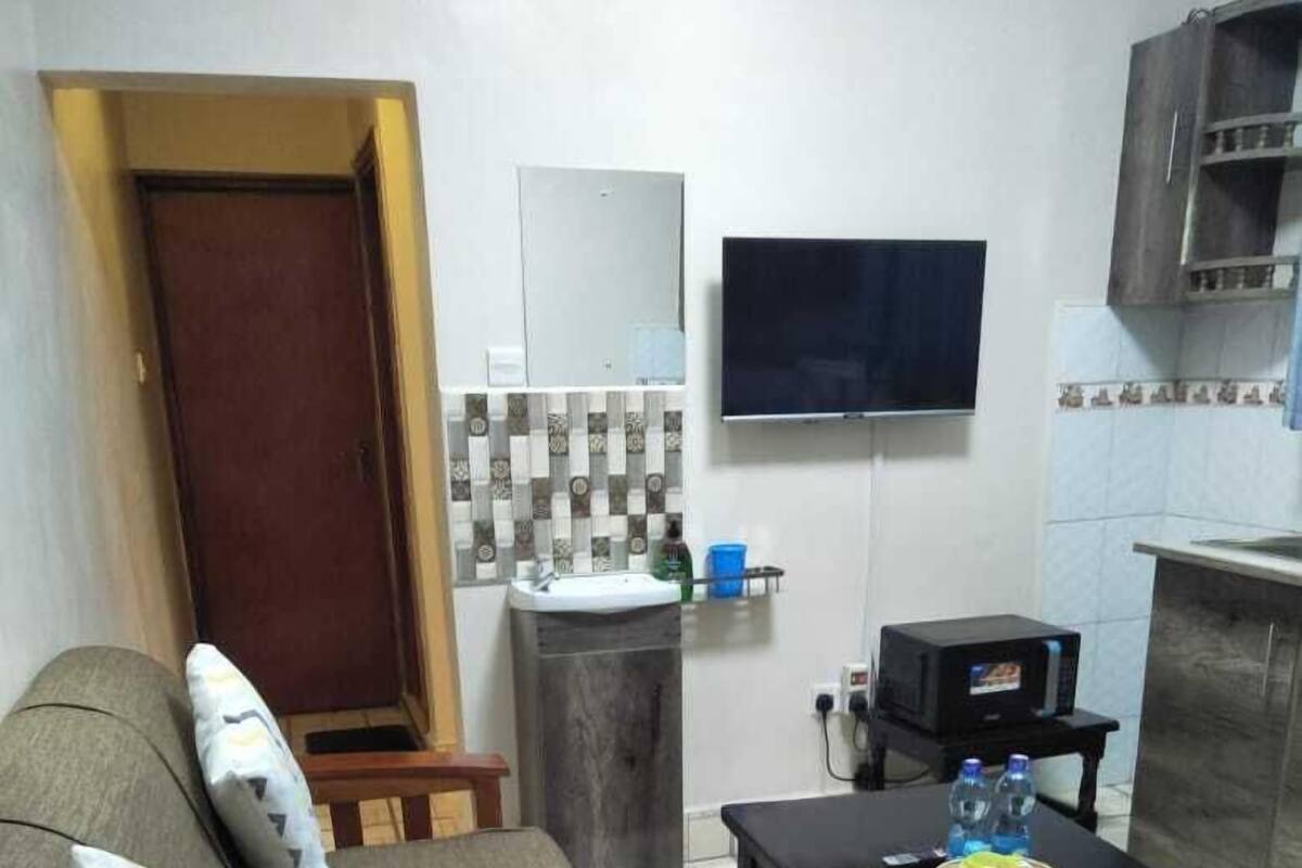 Ein kleiner Wohnraum mit einer Couch, einem Tisch, einer Mikrowelle, einem Spülbecken und einem an der Wand befestigten Fernseher.