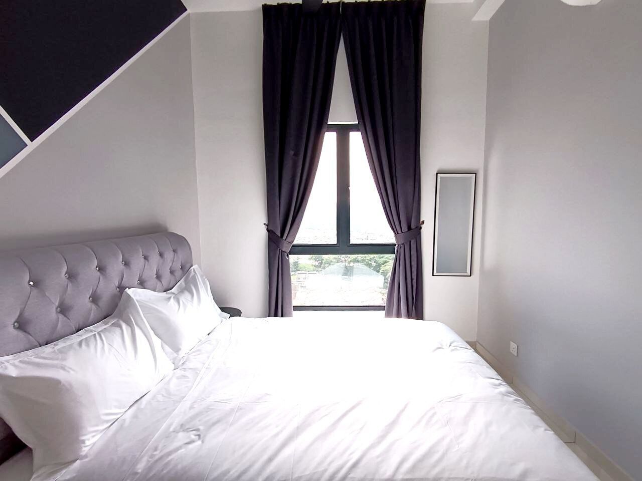 A bedroom with a tufted headboard, a large window with curtains, and a bedside mirror.
