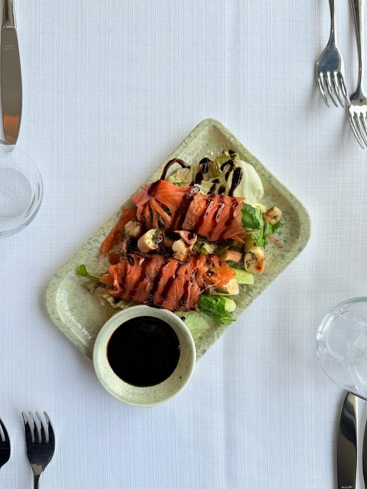 Un plato rectangular con salmón en rodajas, verduras y un pequeño recipiente con salsa, sobre un mantel blanco.