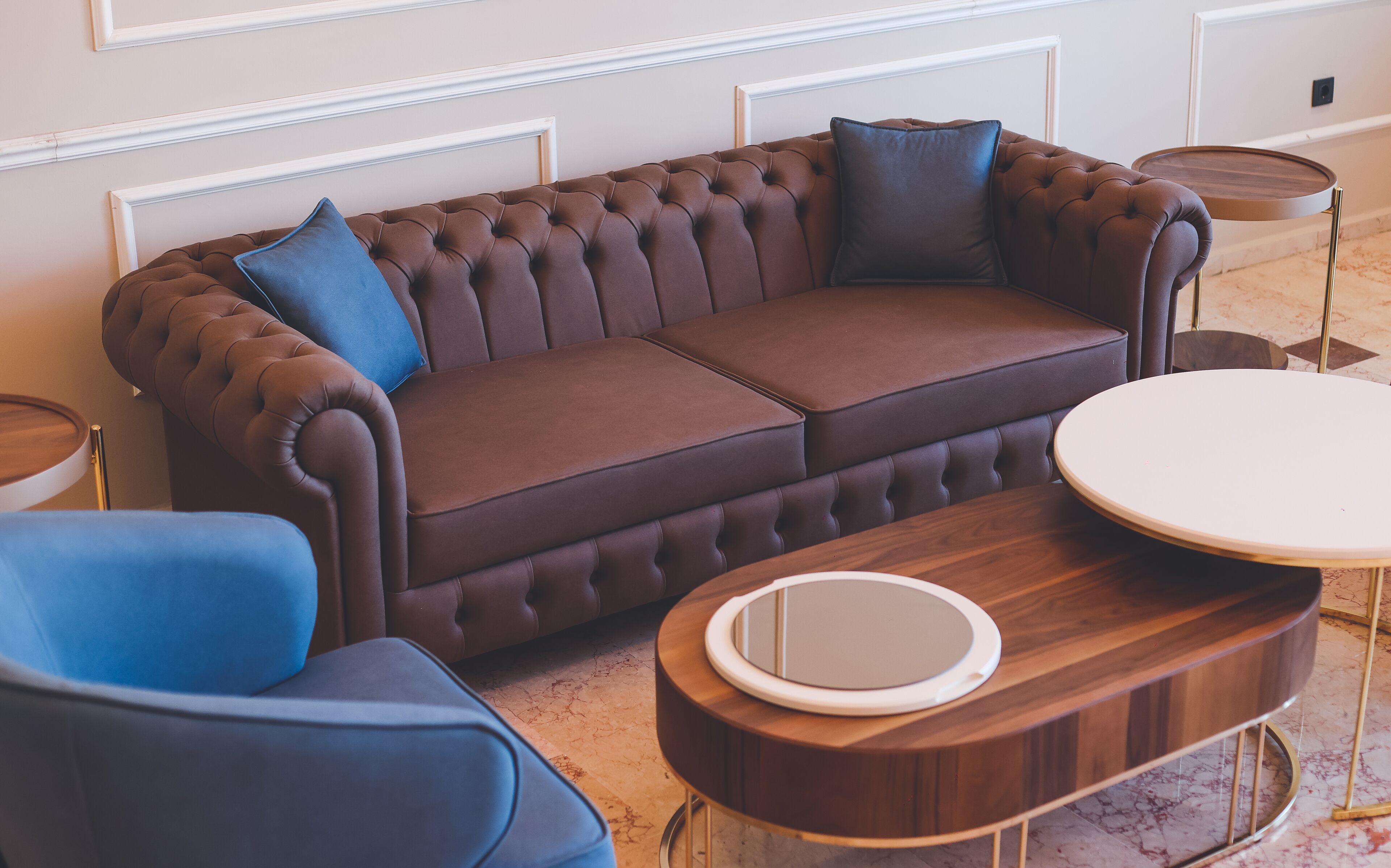 Een bruine sofa met tuftafwerking, twee blauwe kussens, een ronde houten salontafel met een spiegel, en een klein rond tafeltje met een witte blad.