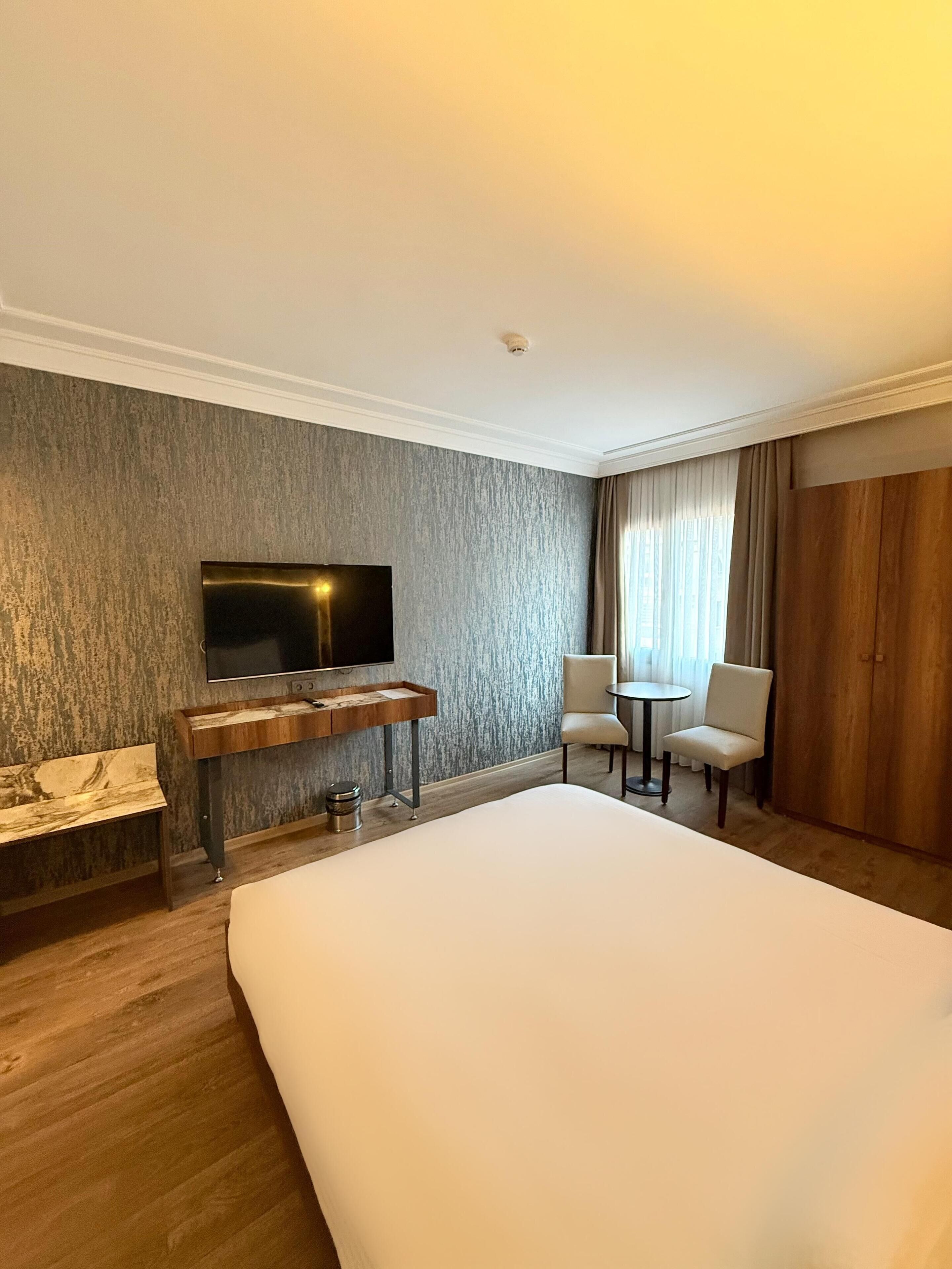 Ein Hotelzimmer mit einem großen Bett, einem flachbildfernseher an einer strukturierten Wand, einem kleinen Holztisch, zwei Stühlen, einem runden Tisch und einem Kleiderschrank.