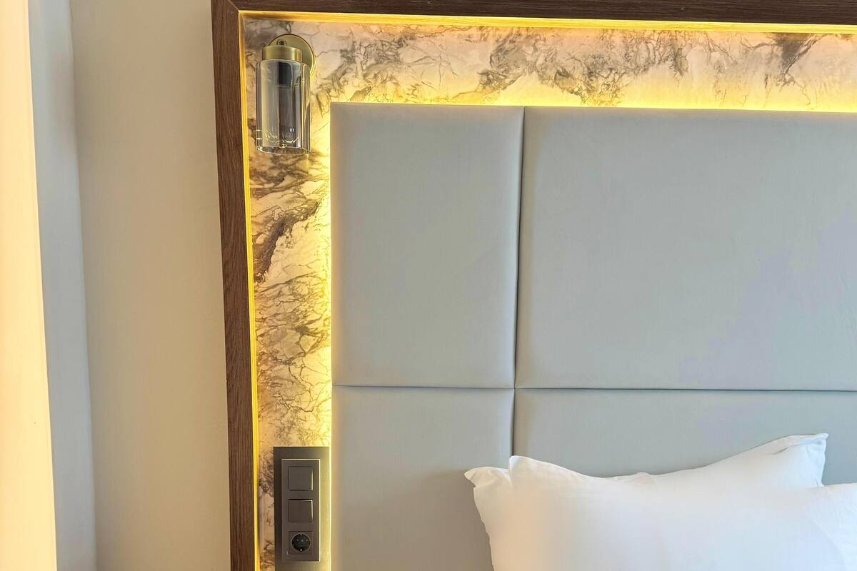 Een net opgemaakt bed met witte lakens en kussens, een houten nachtkastje met een lamp, en een wand met een marmerlook.