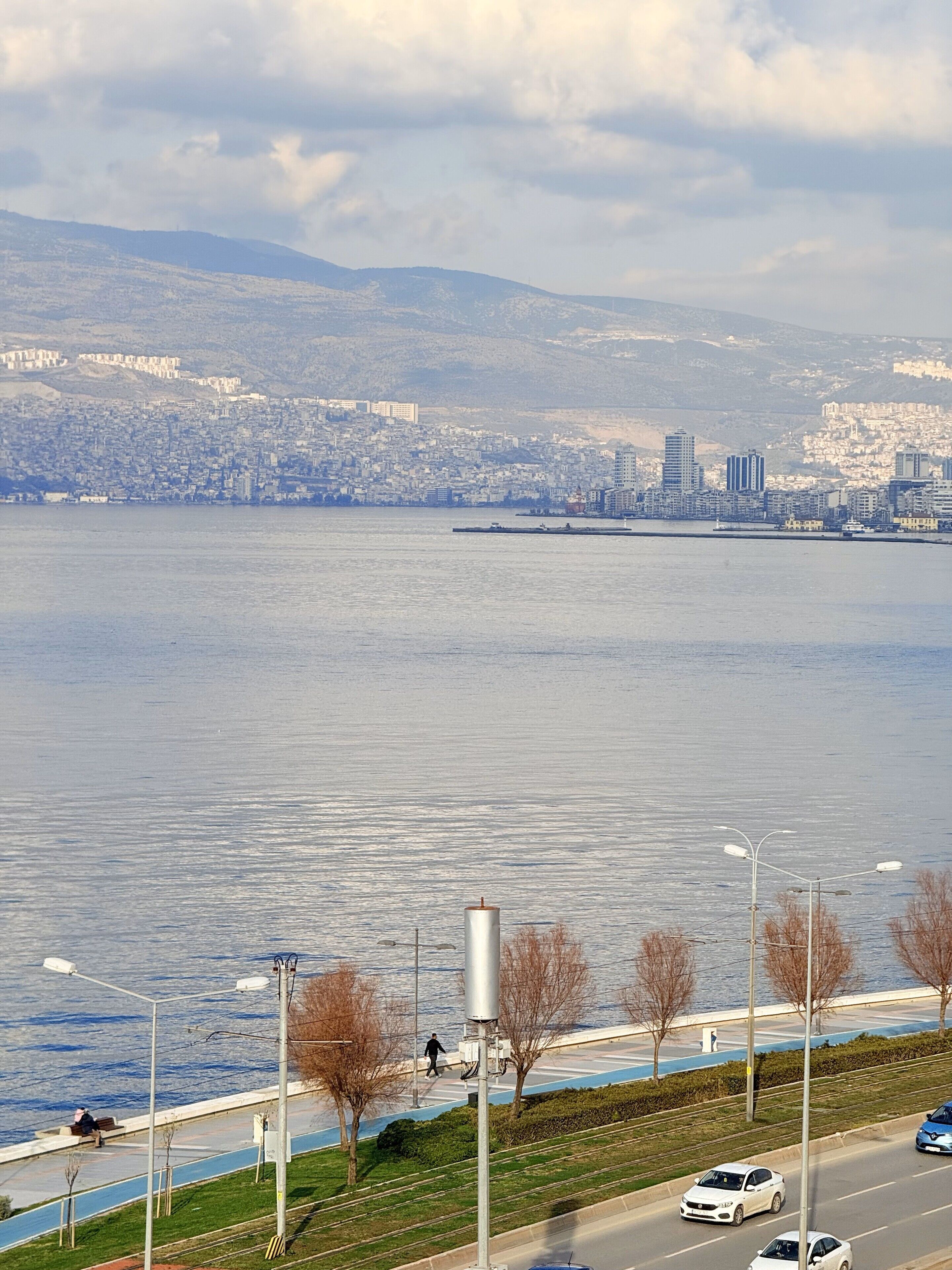 Un paisaje urbano frente al agua, con vistas a la ciudad al otro lado, un camino peatonal y una carretera con coches.