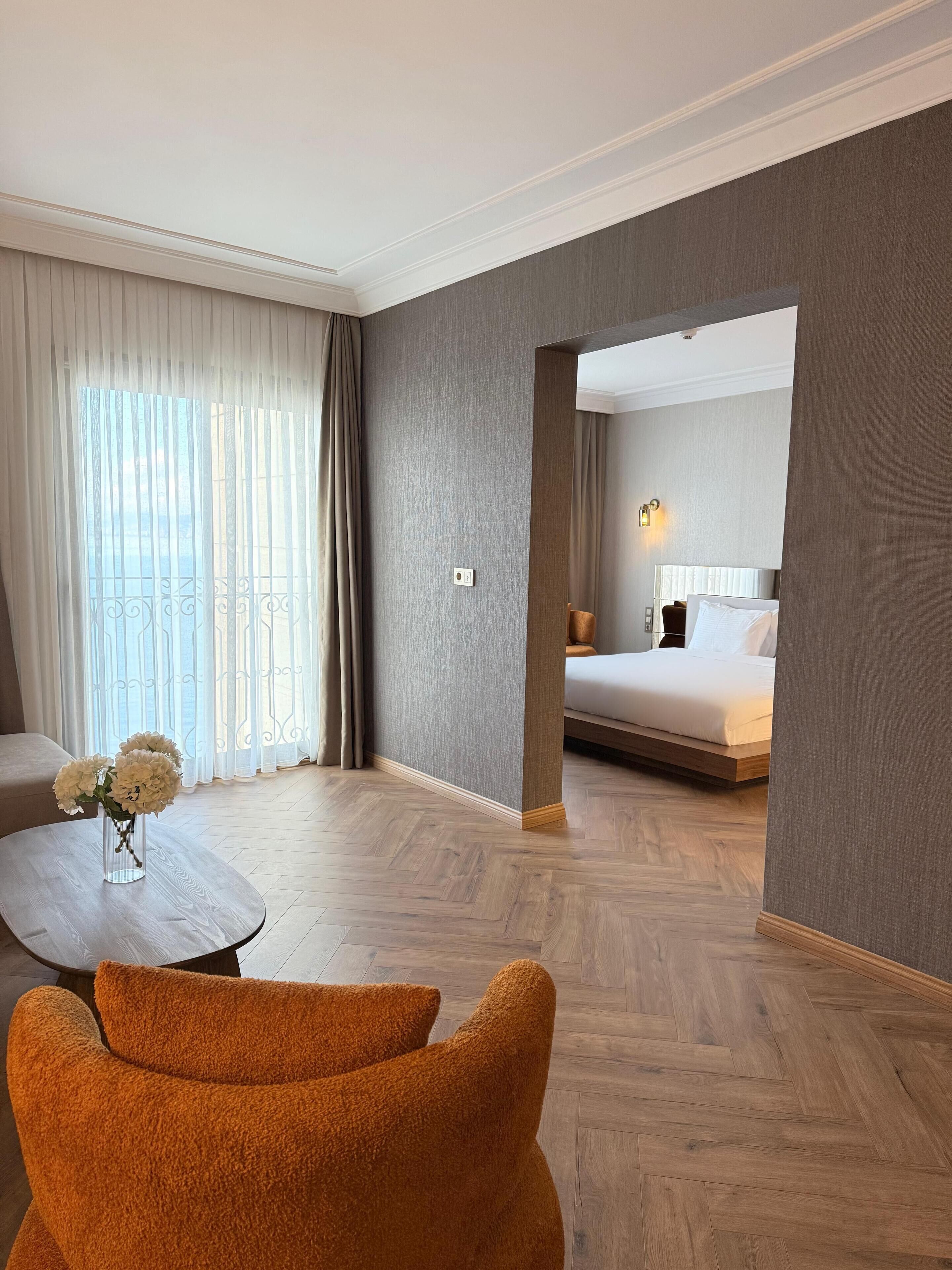 A hotel room with a bed, a seating area with a table and a chair, and a view through an open doorway.