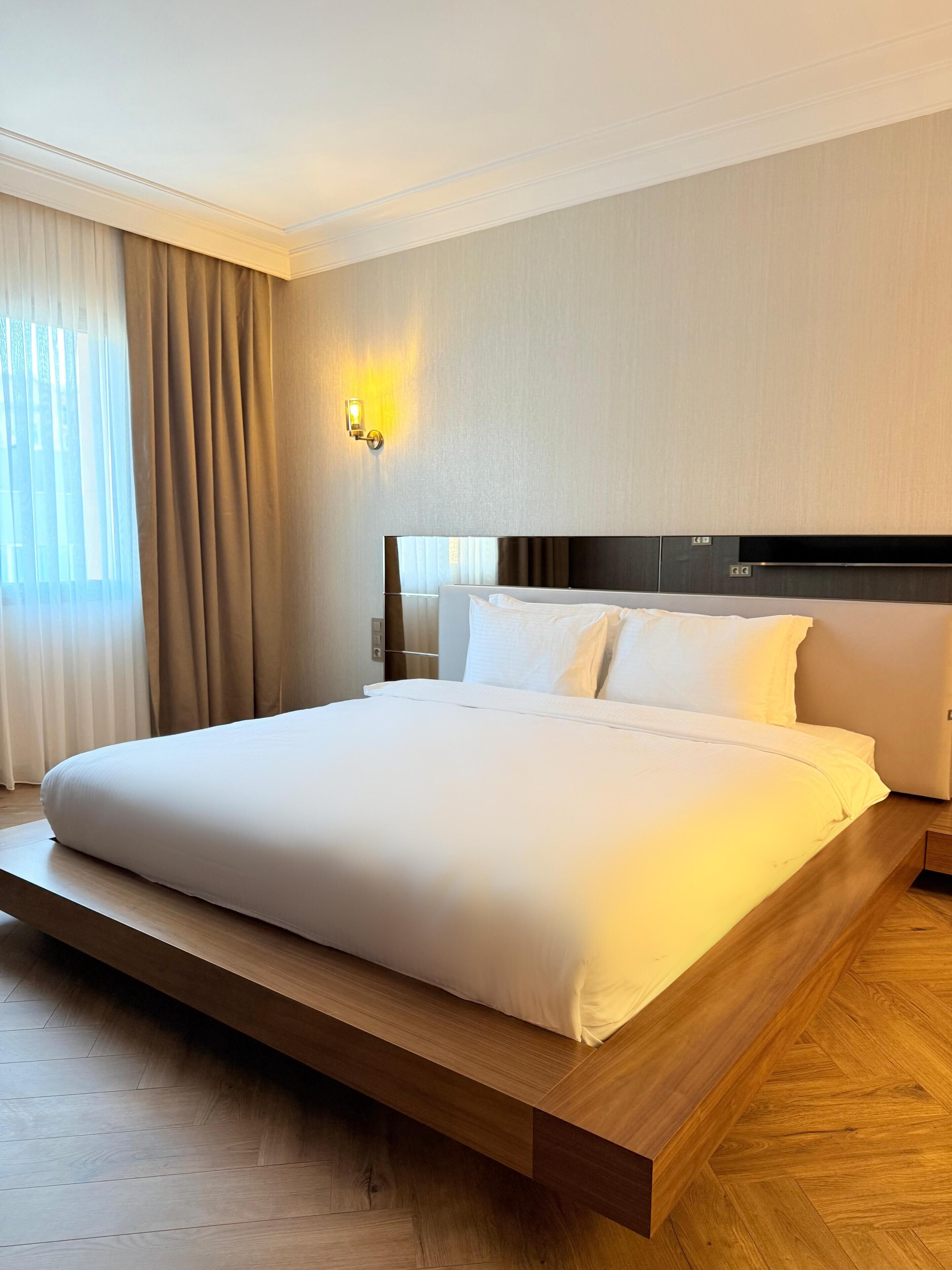 A modern hotel room with a large bed, wooden flooring, and a wall-mounted light fixture.