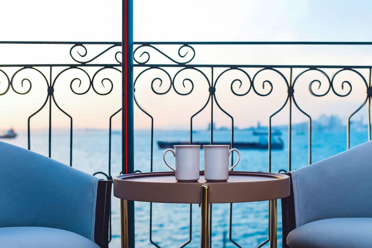 Een balkon met twee witte kopjes op een klein rond tafeltje, uitkijkend op een wateroppervlak met een schip in de verte.