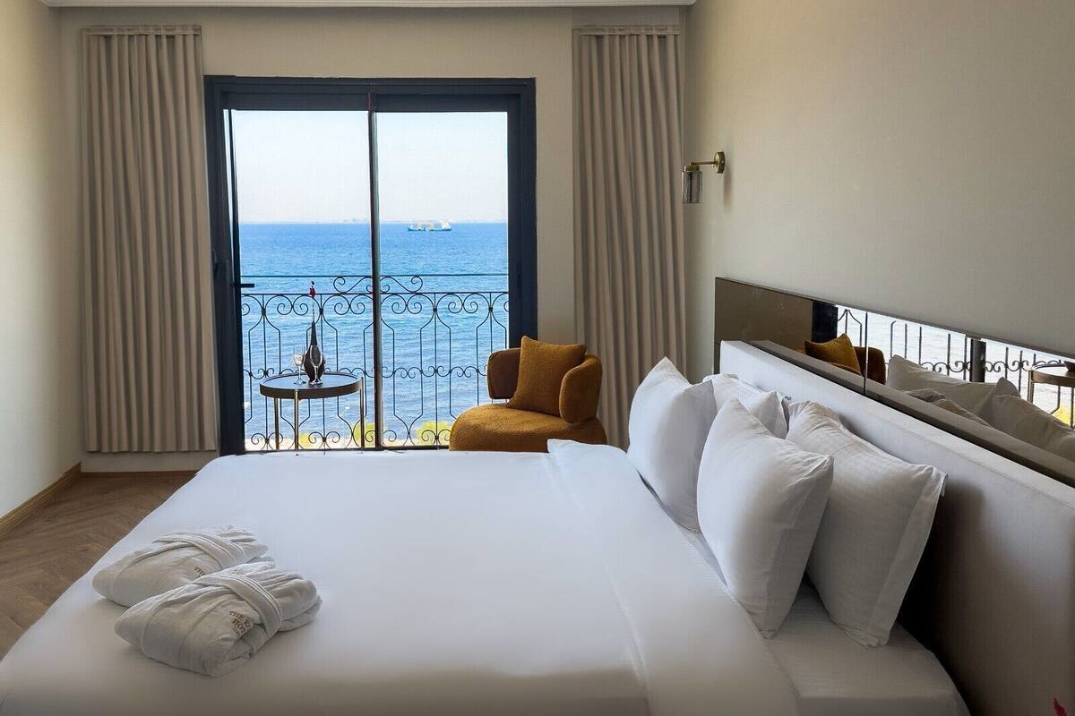Een hotelkamer met een groot bed, een klein tafeltje met een glas, een stoel en uitzicht op de zee vanaf een balkon.