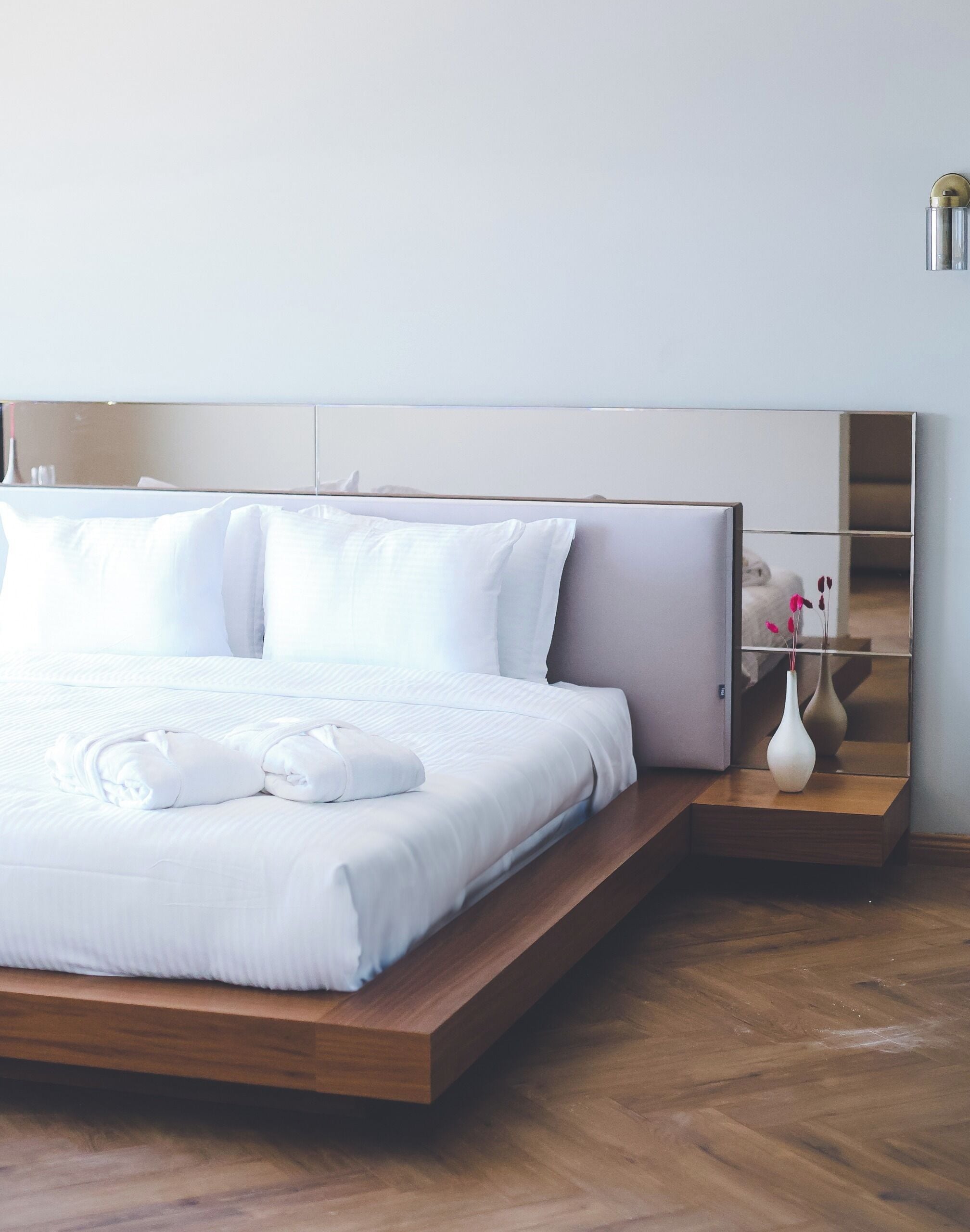 A modern hotel room with a large bed, wooden platform, white bedding, a bedside table with a vase, and a mirror on the wall.
