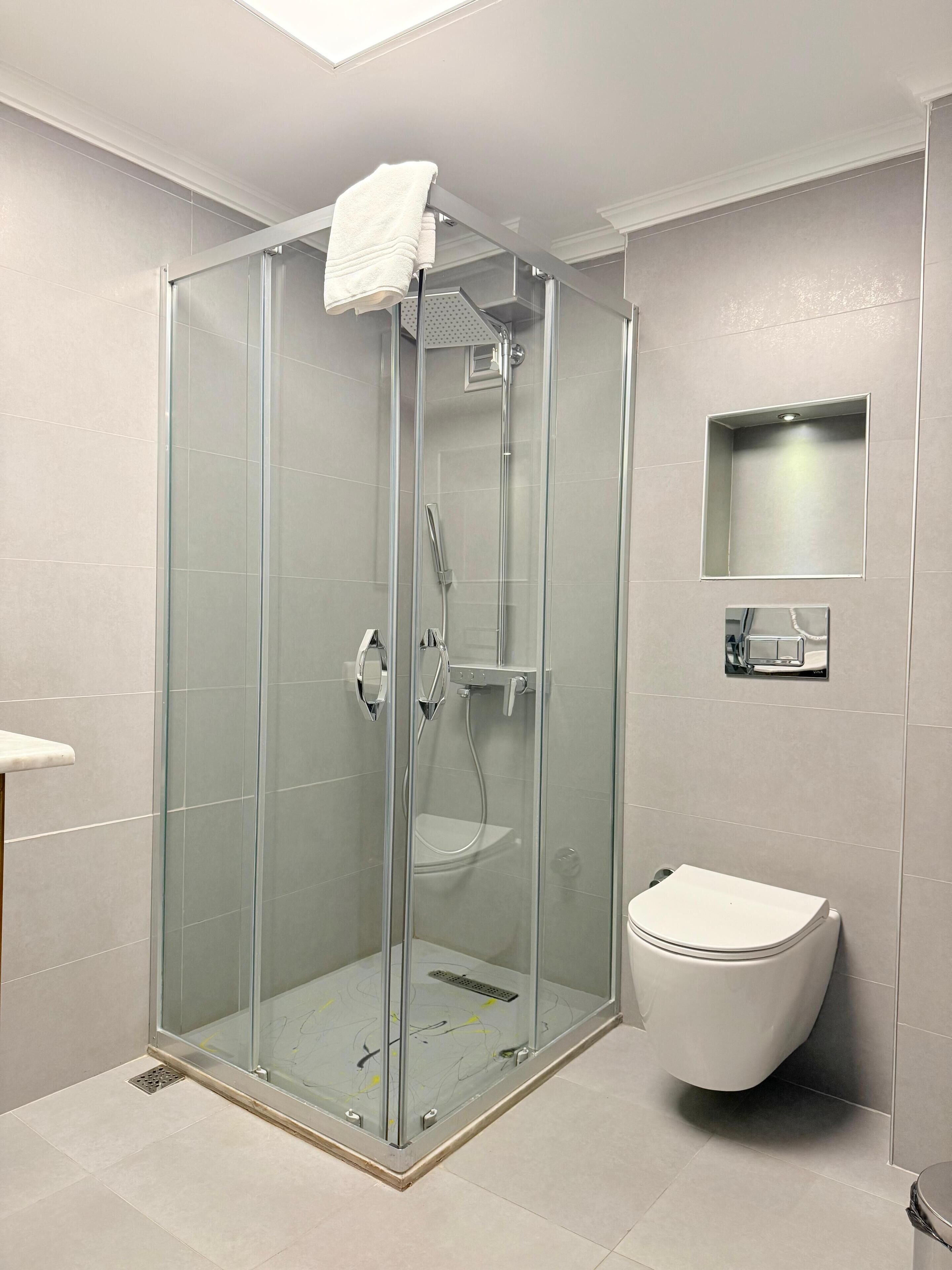 Een moderne badkamer met een douche met glazen wand, een wit toilet en een klein plankje met een spiegel.