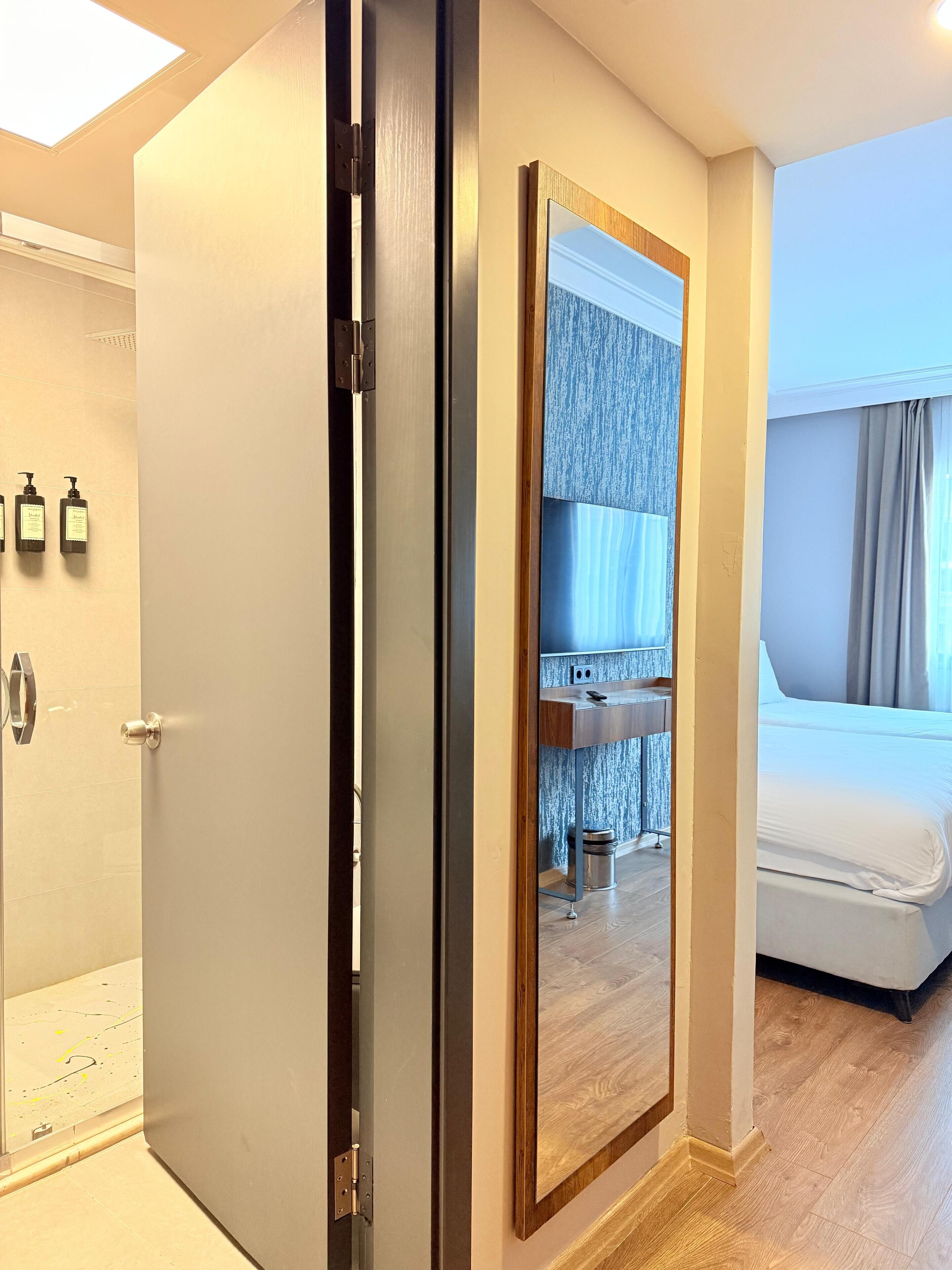 A hotel room with a large mirror, a bed, a bathroom with toiletries, and a view of the city.