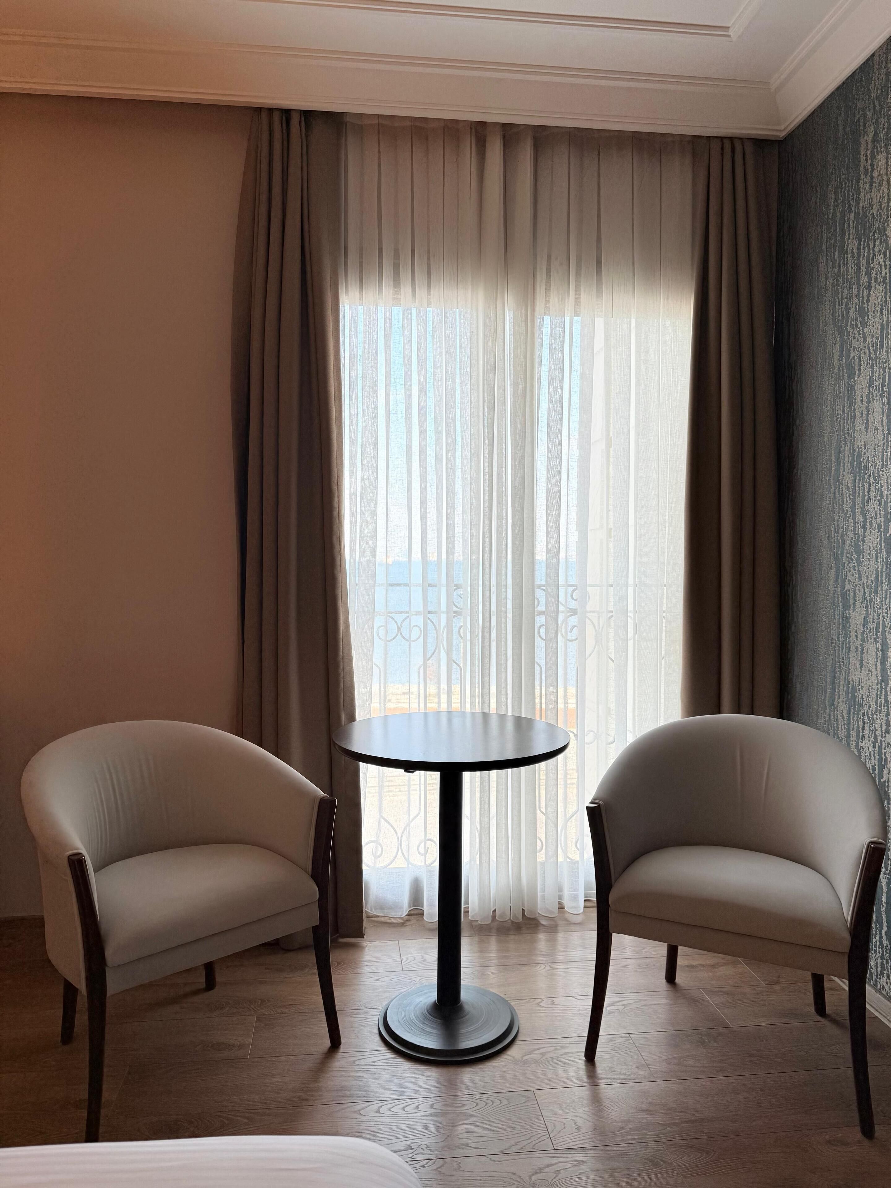 Una habitación con dos sillones y una mesa redonda, un ventanal con cortinas ligeras y vistas al exterior.