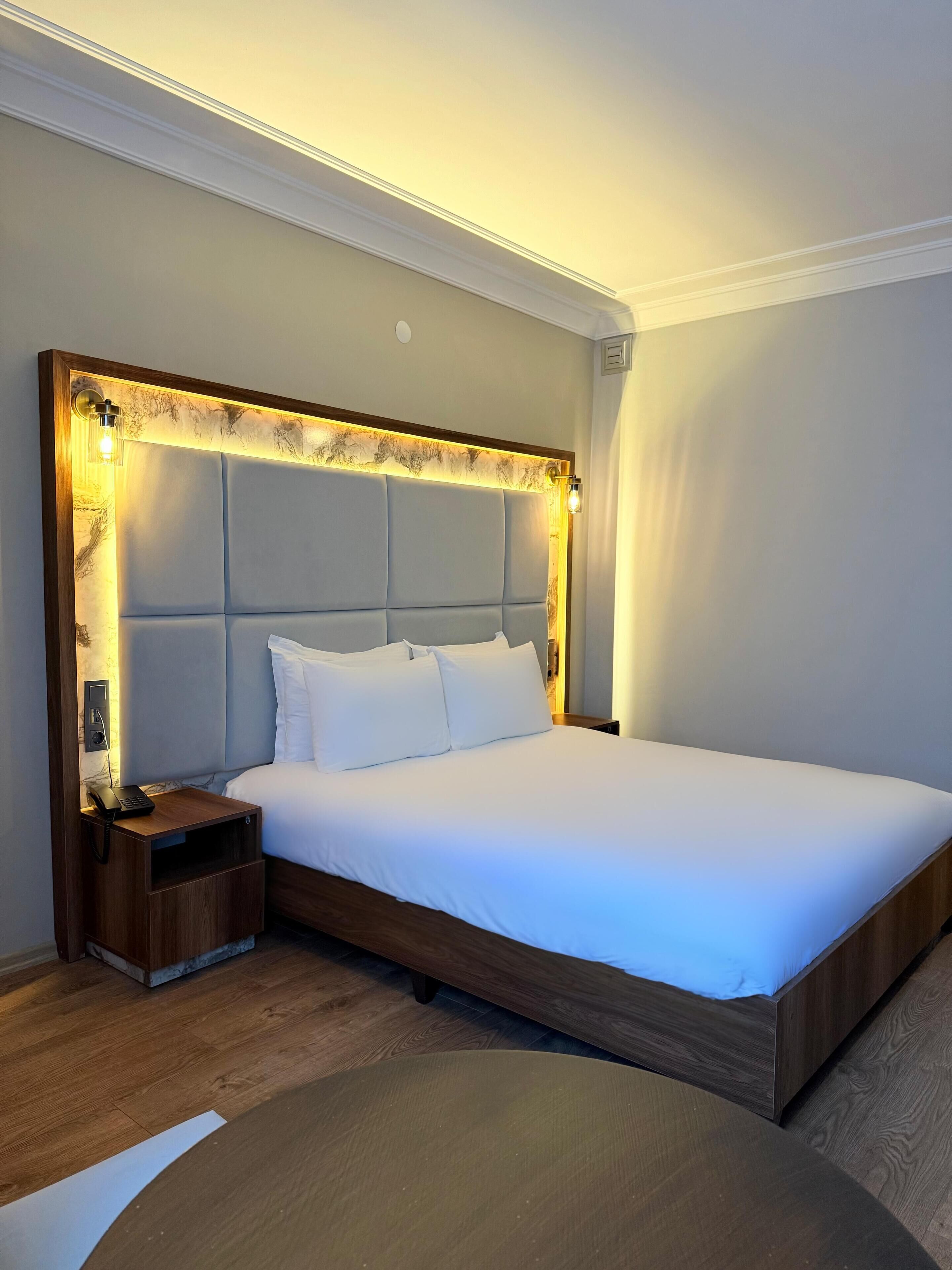 A neatly made bed with a wooden headboard, a bedside table with a lamp, and a small drawer unit.
