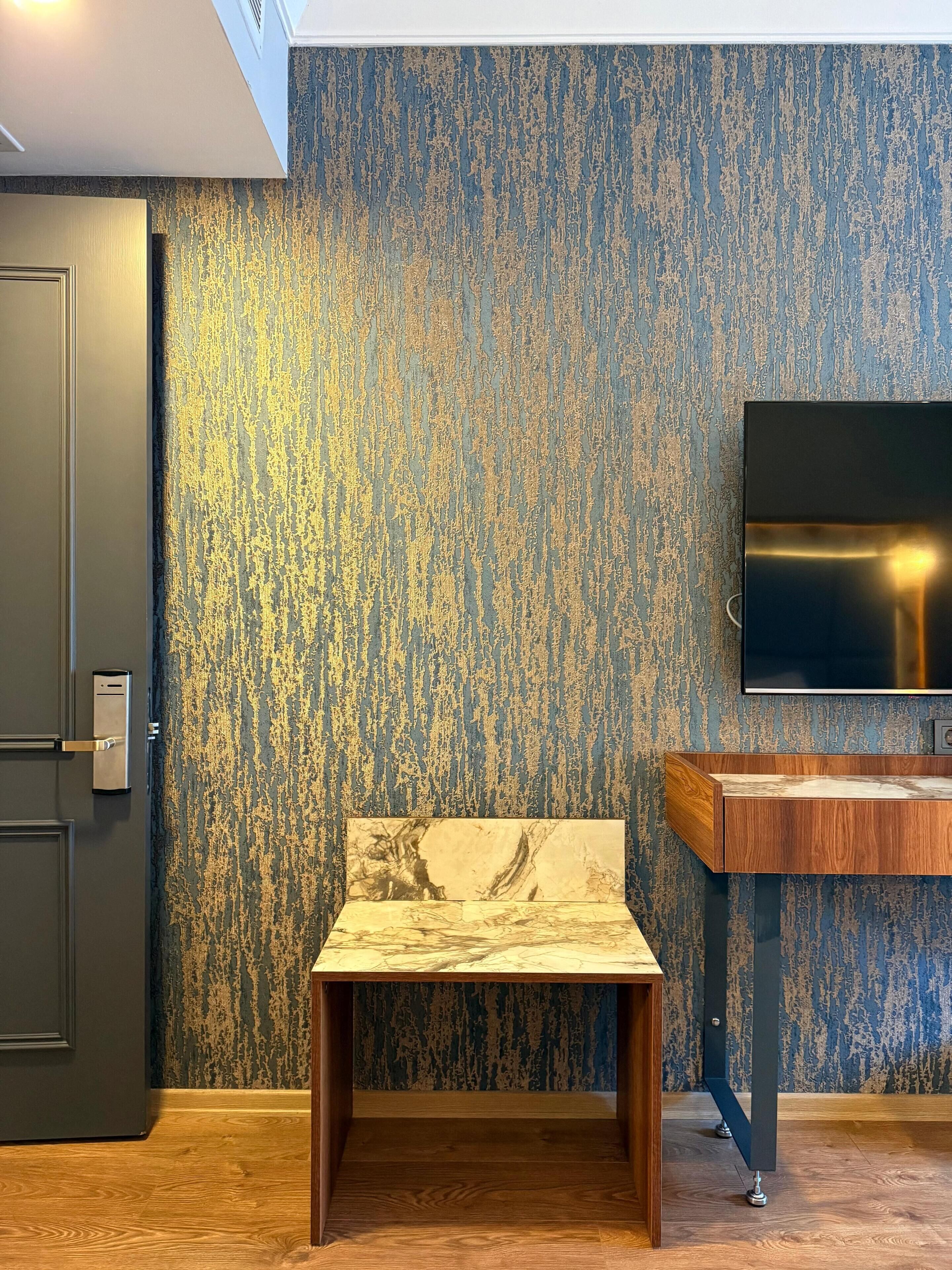 A room with a textured blue wall, a wooden chair, a wooden desk, a flat-screen TV mounted on the wall, and a door with a modern handle.