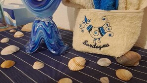 A decorative glass sculpture, a towel with embroidered text, and seashells on a striped fabric. - Summer vacations in Villasimius in a beautiful villa with garden (Sardegna)