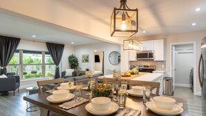 A modern kitchen with white cabinets, a dining area with a wooden table set for a meal, and a view of the outdoors through large windows.