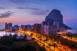 Un paisaje urbano costero al atardecer con una montaña destacada, calles iluminadas y un puerto deportivo.